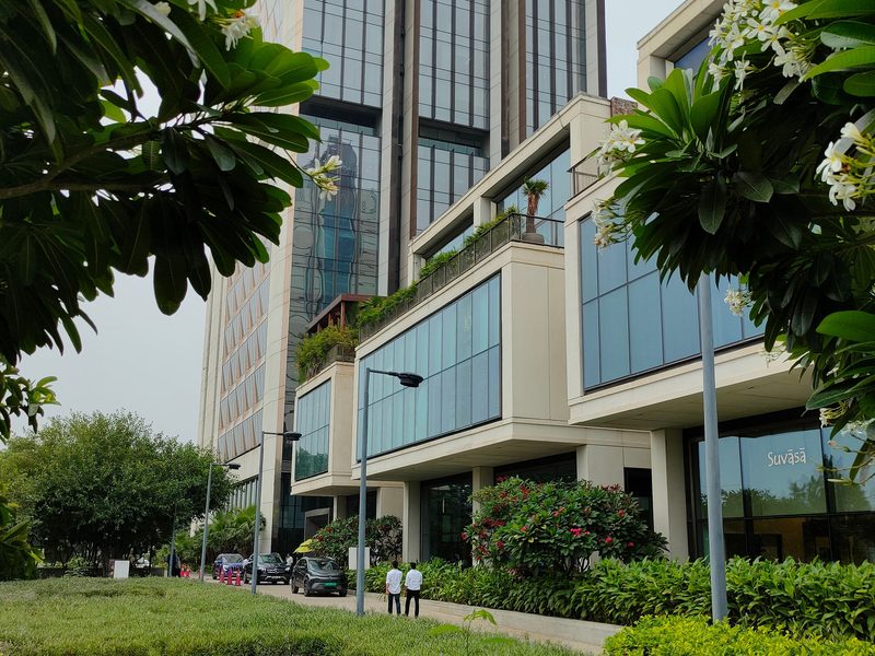 A multi-storey office building behind clipped hedges and trees, with staff crossing the forecourt