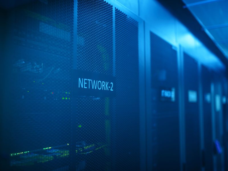 Darkened data centre aisle lined with server and network racks, their status lights glowing blue along the cabinet fronts