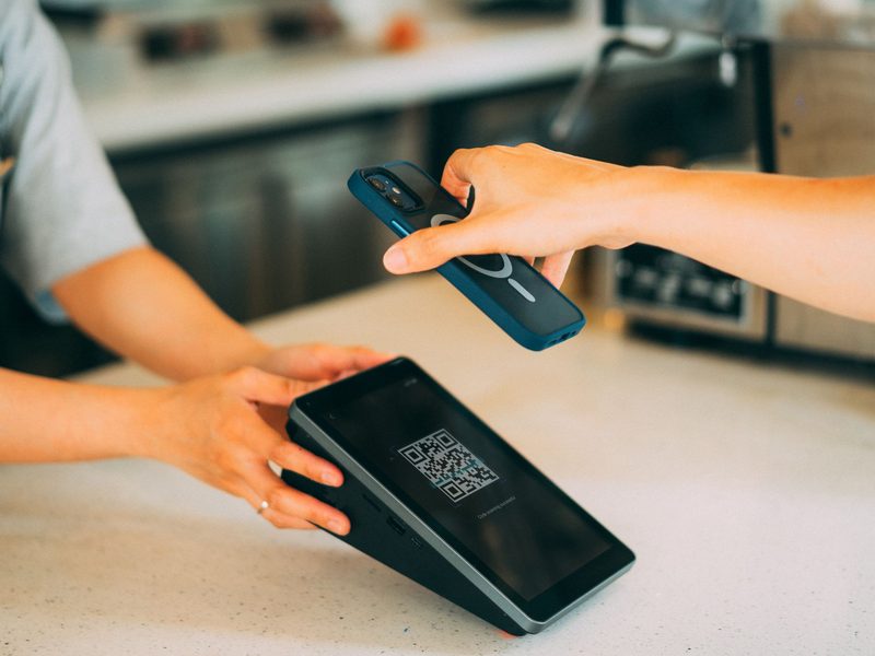 Customer holding a smartphone up to a countertop payment terminal displaying a QR code to complete a contactless transaction
