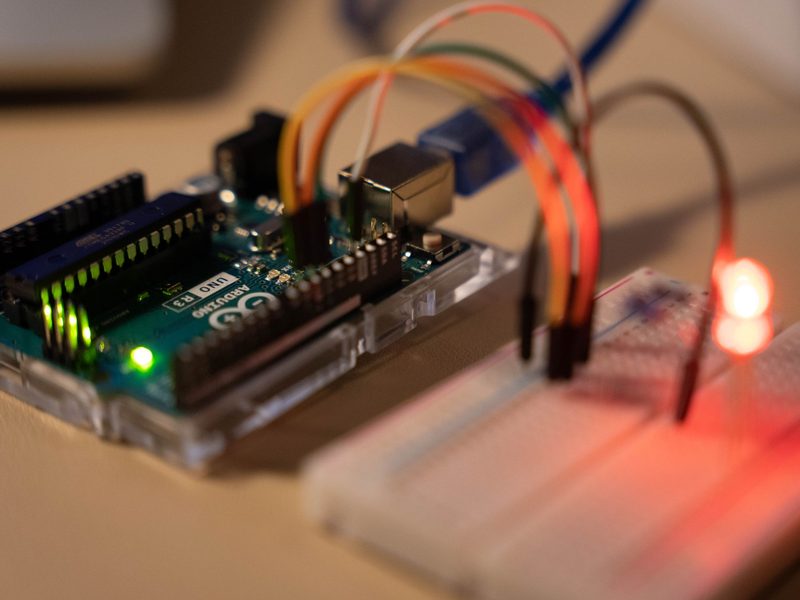 Microcontroller board wired to a breadboard with coloured jumper leads, a red indicator LED lit on the prototype circuit