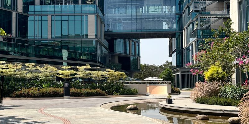 Glass-fronted technology park buildings enclosing a landscaped courtyard with a stone water feature and palms in bright daylight