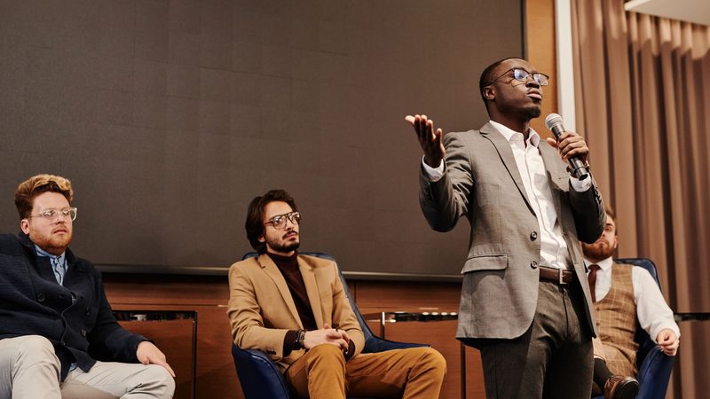 A speaker at a microphone making a point on stage while three fellow panellists listen