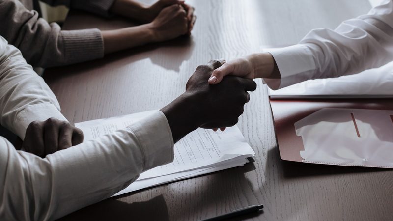 Close view of two people shaking hands across a meeting table over the signed agreement documents in front of them