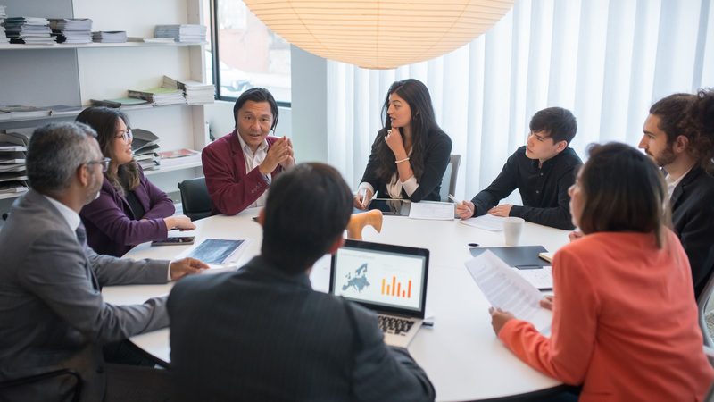 Professionals seated around a boardroom table in discussion, laptops and printed charts spread out in front of them