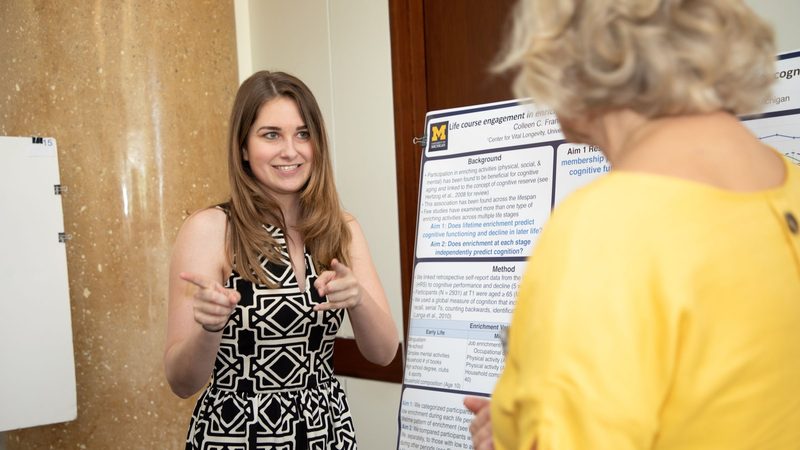 Young woman standing beside her research poster, explaining it with a smile to a visitor at a conference poster session
